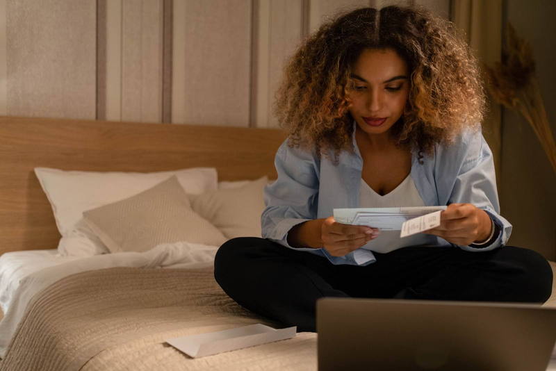 Una mujer sentada en su cama por la noche, leyendo estados de cuenta frente a su computadora.
